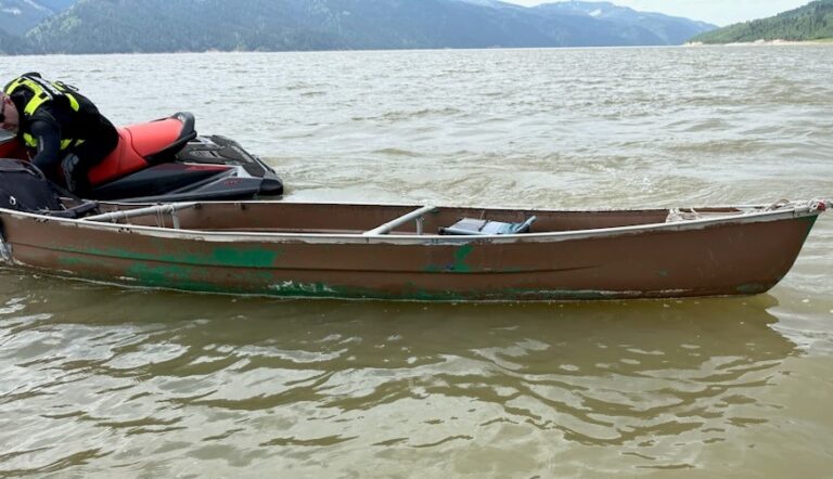 A close-up of an old, brown aluminum canoe floating in a lake beside a jet ski, with mountains and trees visible in the background.
