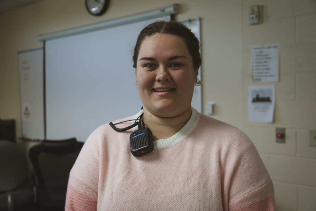 A person with brown hair tied back, wearing a pale pink sweater and a lanyard with a speaker or microphone device, stands smiling in a classroom with a whiteboard and clock in the background.