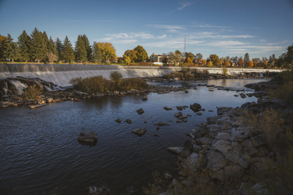 A wide river flows over a low waterfall, surrounded by rocks and autumn trees with colorful leaves under a blue sky. Buildings and a tall tower are visible in the distance.