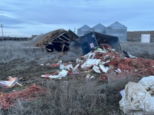 A blue dumpster is tipped over in a field, spilling trash and red debris. A collapsed wooden structure and several metal grain silos are visible in the background under a cloudy sky.