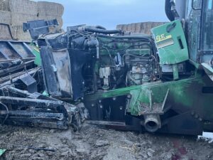 A damaged green tractor with exposed engine parts sits on muddy ground, with hay bales and equipment visible in the background. The front of the tractor is missing panels and appears heavily wrecked.