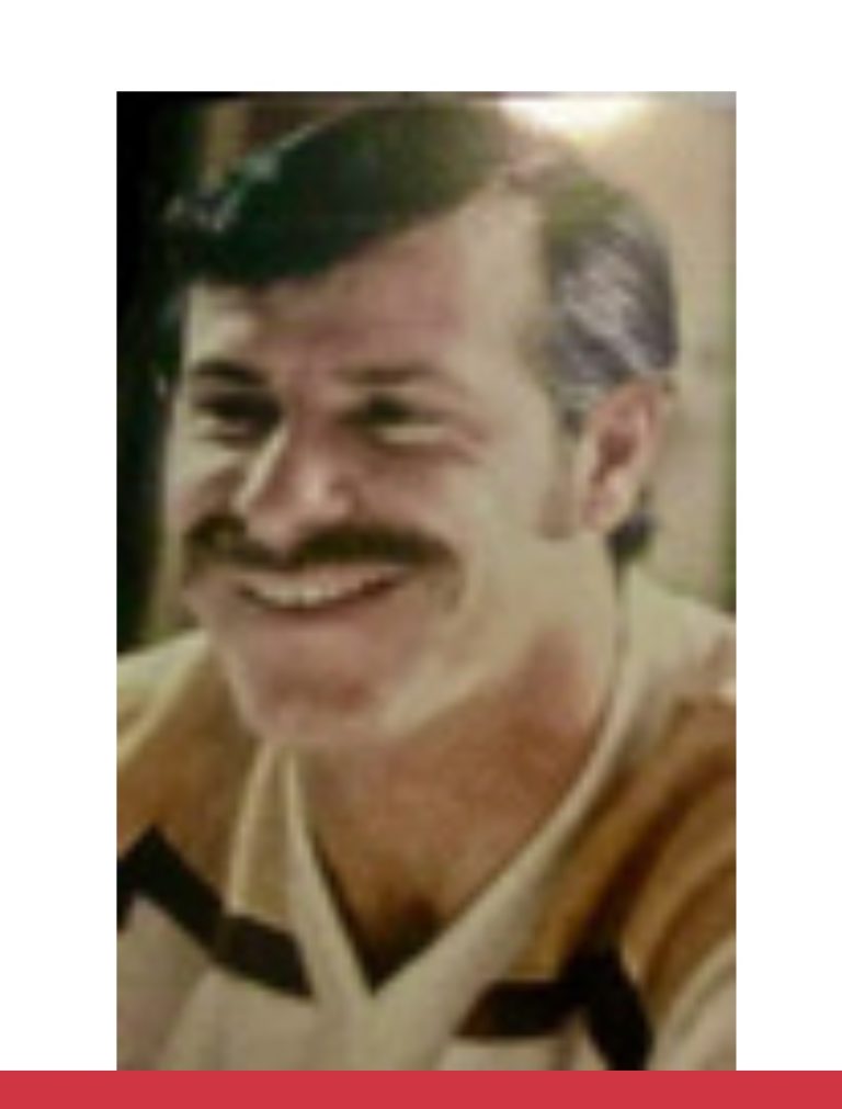 A man with short dark hair and a mustache is smiling. He is wearing a light-colored shirt with dark stripes. The background is softly lit and blurred.
