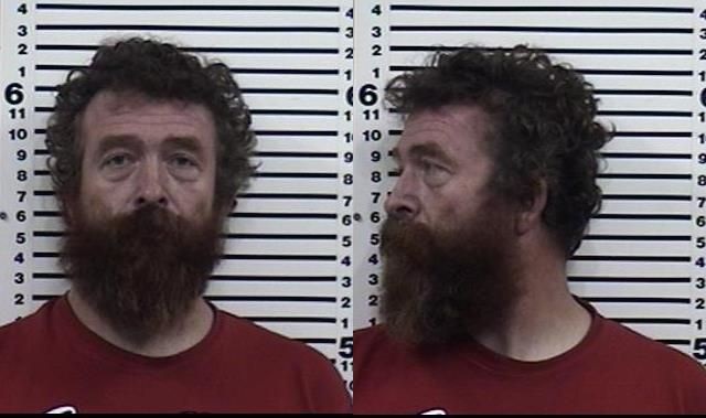A man with curly hair and a thick beard, wearing a red shirt, is shown in front and side mugshot photos against a height chart background.