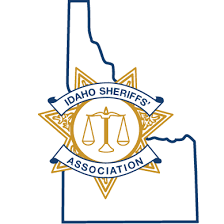Outline of Idaho state with a gold sheriff’s badge over it. The badge has a justice scale in the center and a blue ribbon that reads “Idaho Sheriffs’ Association.”.