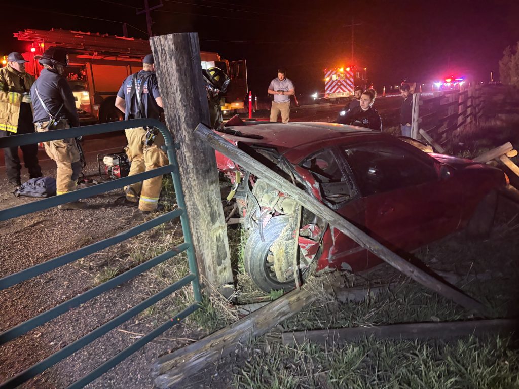 A red car crashed into a wooden fence at night, with significant rear damage. Firefighters and emergency personnel are at the scene, and emergency vehicles with flashing lights are visible in the background.