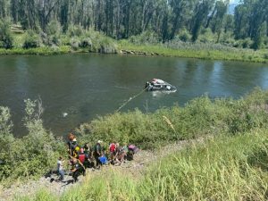 A group of people stands on a grassy riverbank while a partially submerged vehicle is pulled from the river with a rope. Rescue workers and onlookers are gathered near the water’s edge. Trees line the background.