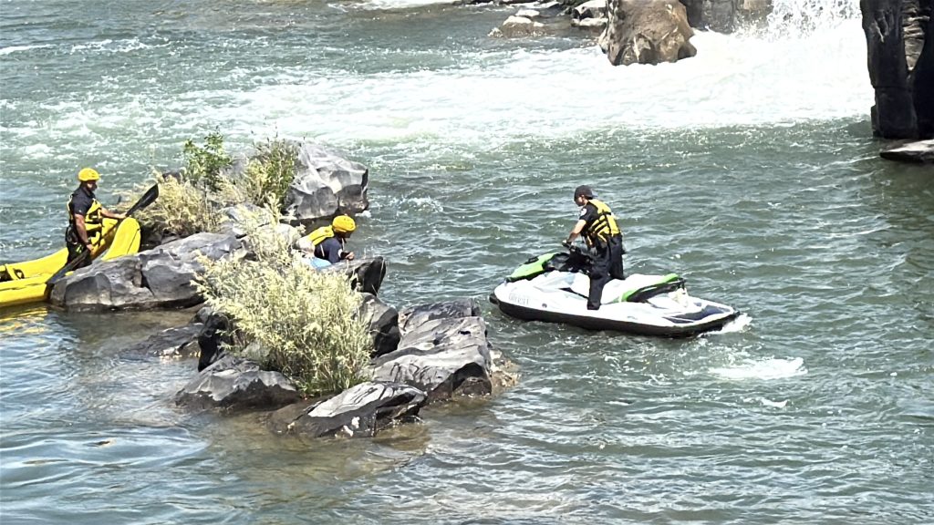 People wearing yellow helmets and life jackets are rafting and riding a jet ski near rocks in a fast-moving river with white water and small rapids.