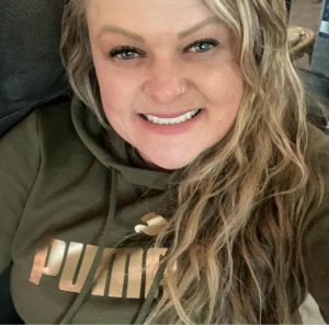 A smiling person with long wavy blonde hair wearing an olive green Puma hoodie, sitting indoors.