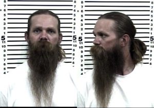 Mugshot of a man with a long brown beard and hair tied back, wearing a plain white shirt, shown from the front and side against a height chart background.