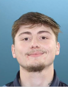 A young man with short light brown hair, a small goatee, and a faint mustache smiles slightly in front of a blue background. He is wearing a dark collared shirt.