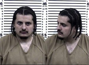 A mugshot of a man with braided hair wearing a tan prison uniform, shown from the front and side against a height chart background.