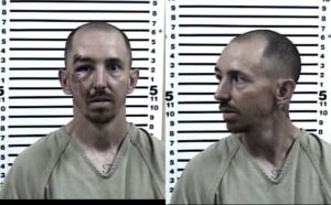 Two mugshot photos of a man wearing a beige prison uniform, shown facing forward and in profile. He has short hair, a goatee, facial injuries, and a chest tattoo. A height chart is visible in the background.