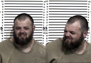 Two mugshots of a bearded man in a tan shirt, facing forward in one and turning to the side in the other, with a height measurement chart in the background.