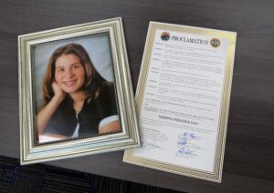A framed photo of a smiling woman is next to an official proclamation document titled “Missing Person’s Day,” both placed on a gray table.